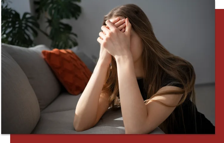 Woman with long hair sitting on a couch, covering face in hands, feeling overwhelmed and sad in a sunlit room